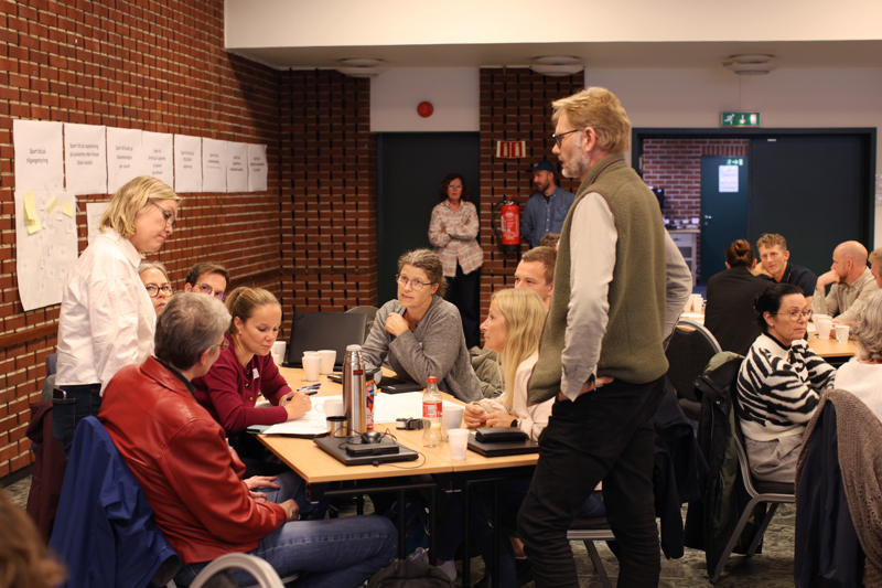 Flere personer sitter rundt et bord og diskuterer. Noen står. 