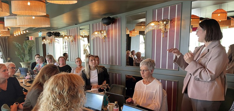 Kristine Skjøthaug står i sofaen og snakker til mange som sitter rundt bordet.