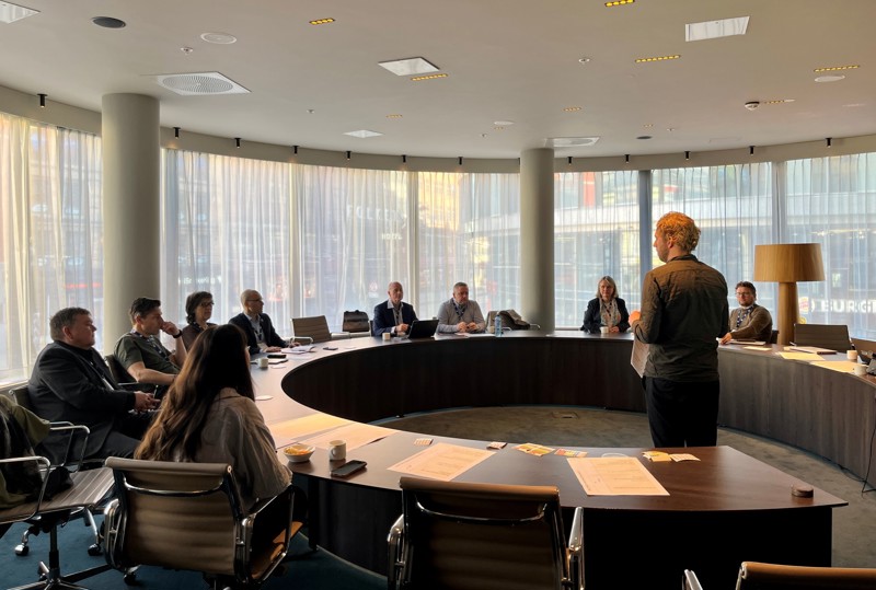 Vi ser et stort ovalt bord i møterommet hvor det sitter folk rundt, og Eirik Haglund står i midten. 