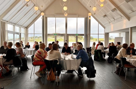 Bildet viser hele lokalet hvor møtet ble holdt. Vi ser mange runde bord, hvor det sitter folk rundt som snakker med hverandre.