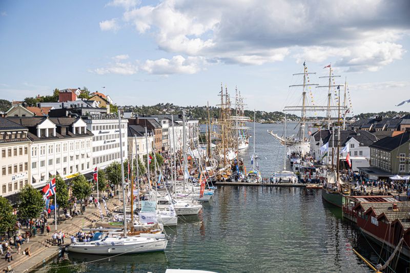 Rundt 1700 arrangementer og 200 stands fyller Arendal sentrum i fem dager i august hvert år. Fotoet viser havnen og sentrum av Arendal. Det ligger mange seilbåter til kai og vi ser folk som går rundt havnen, blant boder og blomsterbed.