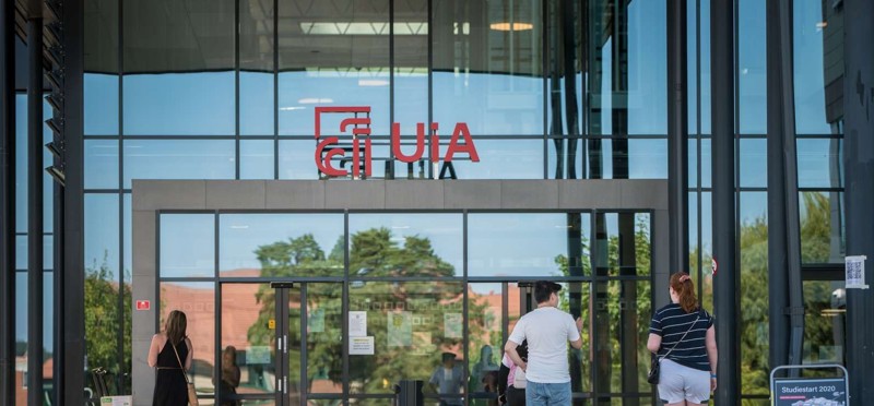 Bildet viser hovedinngangen på Universitetet i Agder. Store glassflater, og vi ser noen studenter som går mot inngangen.